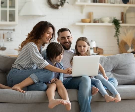 A mother and father with their two children sitting on a couch while using a laptop